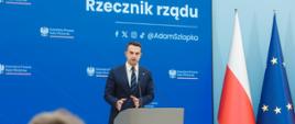 Rzecznik rządu, Minister Adam Szłapka podczas konferencji prasowej 24.06.2025 r. W tle ekran Kancelarii Prezesa Rady Ministrów oraz media społecznościowe rzecznika rządu. Obok flagi Polski i Unii Europejskiej.