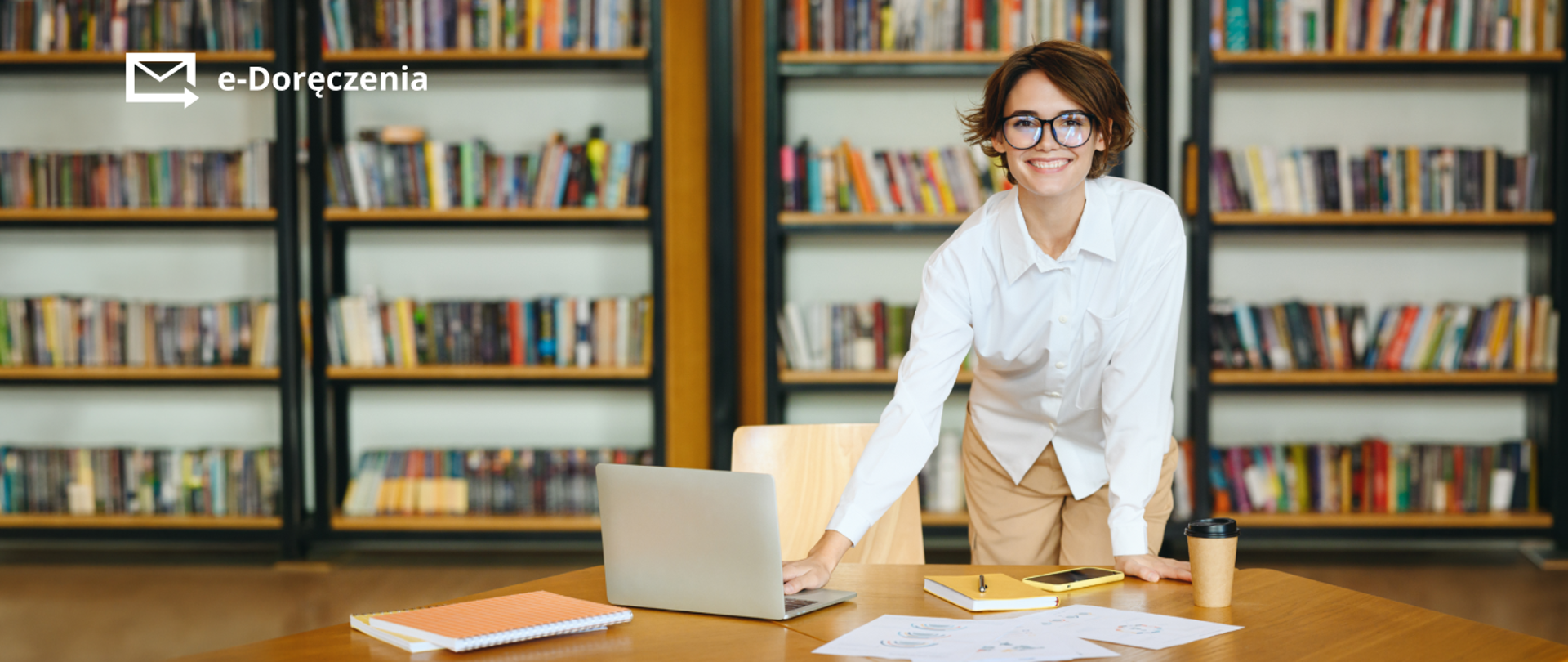 Uśmiechnięta kobieta z otwartym laptopem na biurku, w tle regały z książkami. 