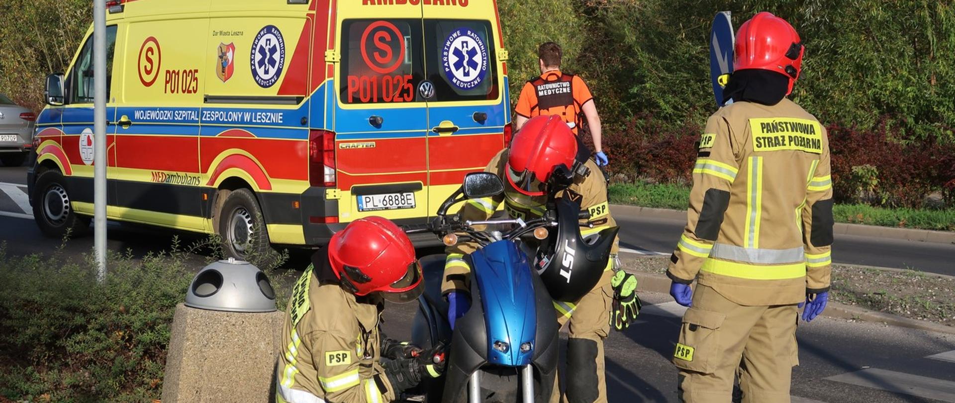 Strażacy prowadzą działania przy skuterze. W tle widoczna karetka pogotowia i ratownik medyczny.
