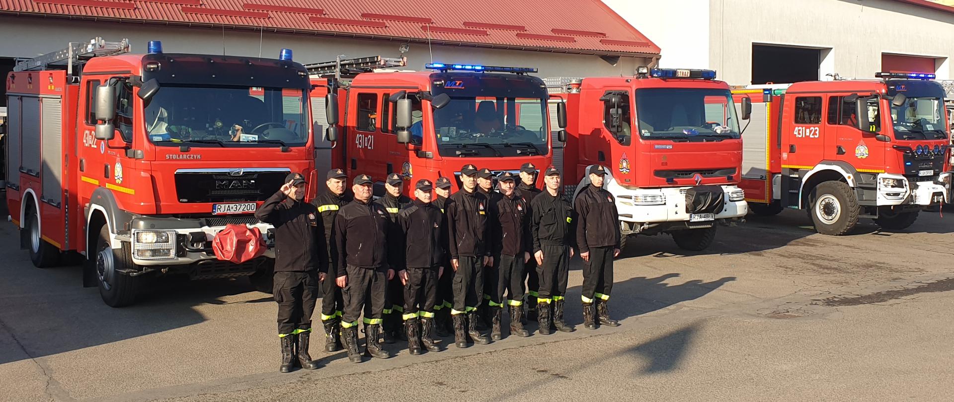 Na zdjęciu widać strażaków stojących na baczność w dwuszeregu, ubranych w ubranie koszarowe. Dowódca pododdziału oddaje honor salutując. Za strażakami widoczne są pojazdy gaśnicze.