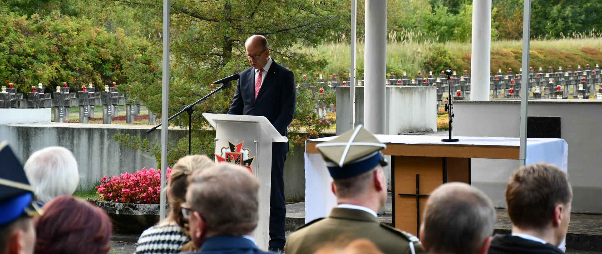 Zdjęcie przedstawia moment odczytania listu przez ubranego w ciemny garnitur, białą koszulę i czerwony krawat mężczyznę. Przed nim znajdują się uczestnicy uroczystości. 