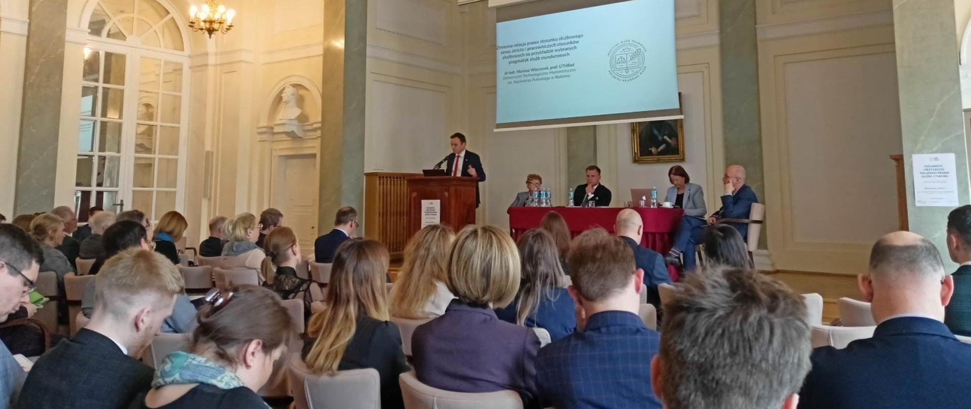 W eleganckiej sali konferencyjnej siedzi kilkadziesiąt osób. Przy mównicy mężczyzna z garniturze, na wyświetlaczu tytuł panelu.
