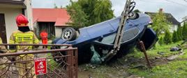 Uszkodzony pojazd osobowy marki BMW na boku po uderzeniu w betonowy słup energetyczny. Obok strażak w umundurowaniu bojowym. 