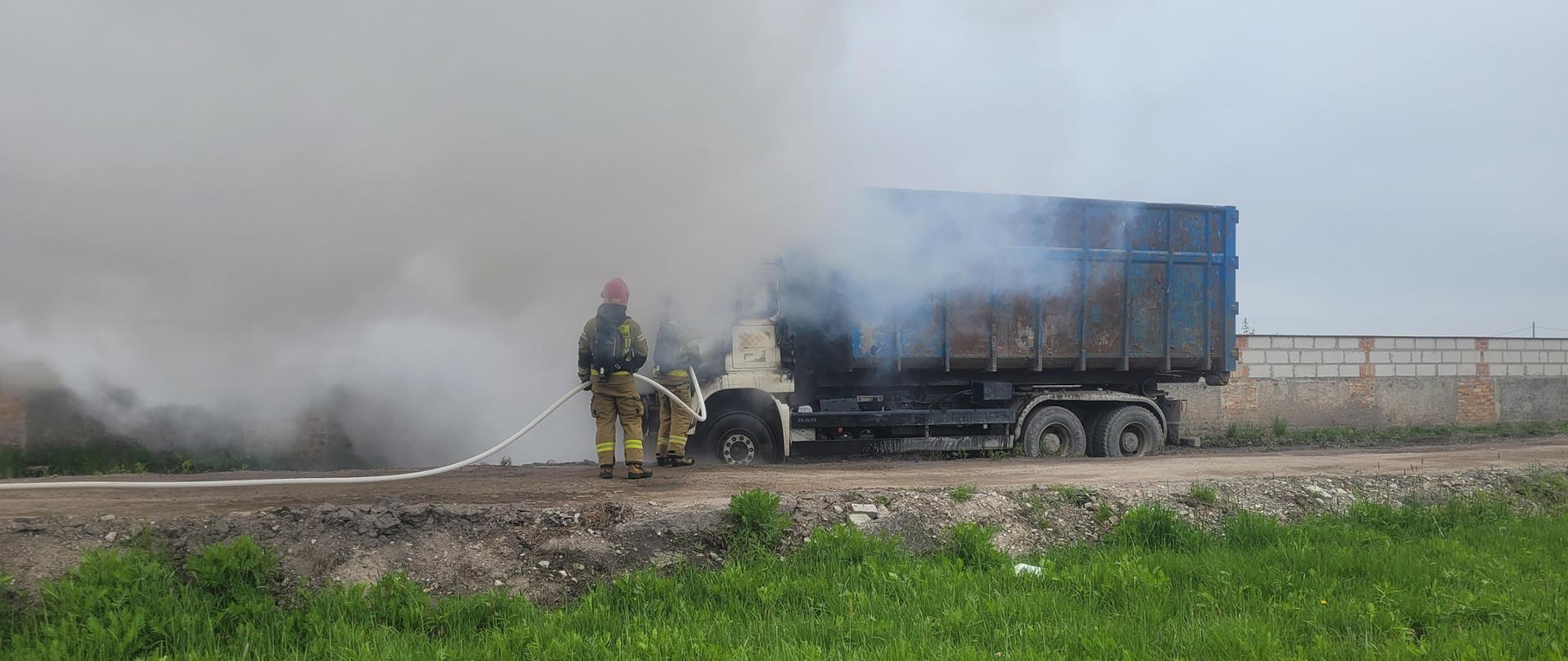 Zdjęcia przedstawiają działania gaśnicze podczas pożaru samochodu ciężarowego. Na fotografii widoczni są strażacy w ubraniach specjalnych oraz mają założone aparaty ochrony dróg oddechowych. Pożar rozwija się w przestrzeni kabiny pojazdu. Samochód ustawiony jest na drodze gruntowej.