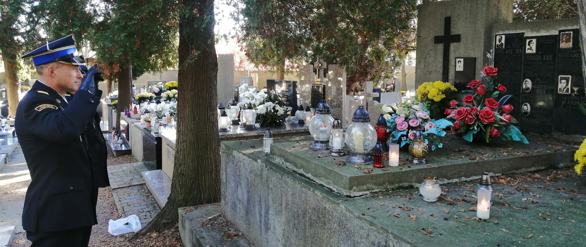 Zdjęcie przedstawia strażaków z Państwowej Straży Pożarnej w mundurach wyjściowych na cmentarzu komunalnym w Bochni. Strażacy stoją przed grobem i oddają honor. W tle znajdują się inne groby oraz drzewa