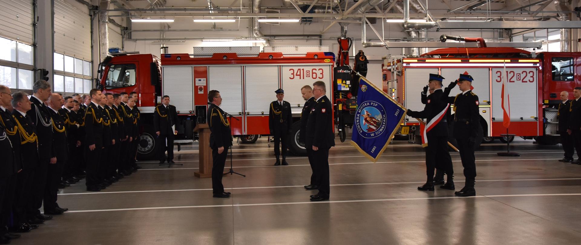 Na garażu Komendy stoją samochody pożarnicze, funkcjonariusze z gośćmi. na planie głównym sztandar Komendy Miejskiej PSP