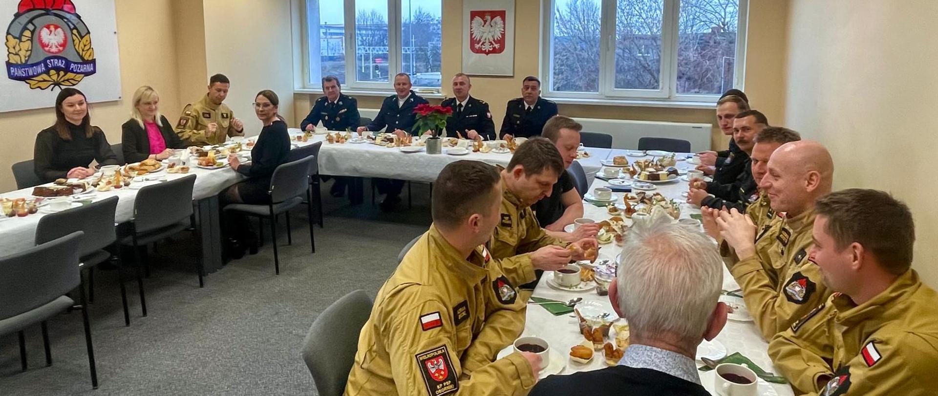 Grupa strażaków Państwowej Straży Pożarnej oraz zaproszonych gości siedzących przy wspólnych stołach podczas uroczystego spotkania w sali konferencyjnej. Na stołach znajdują się ciasta i napoje, a na ścianie widoczny jest emblemat PSP oraz godło Polski.