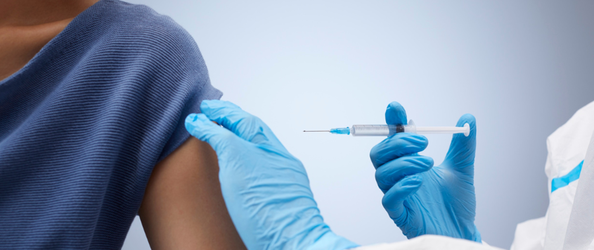 Pielęgniarka w jednorazowych rękawiczkach podaje zastrzyk w ramię osoby siedzącej na krześle. Pacjent odsłania ramię, a medyk uważnie wykonuje szczepienie w sterylnych warunkach.