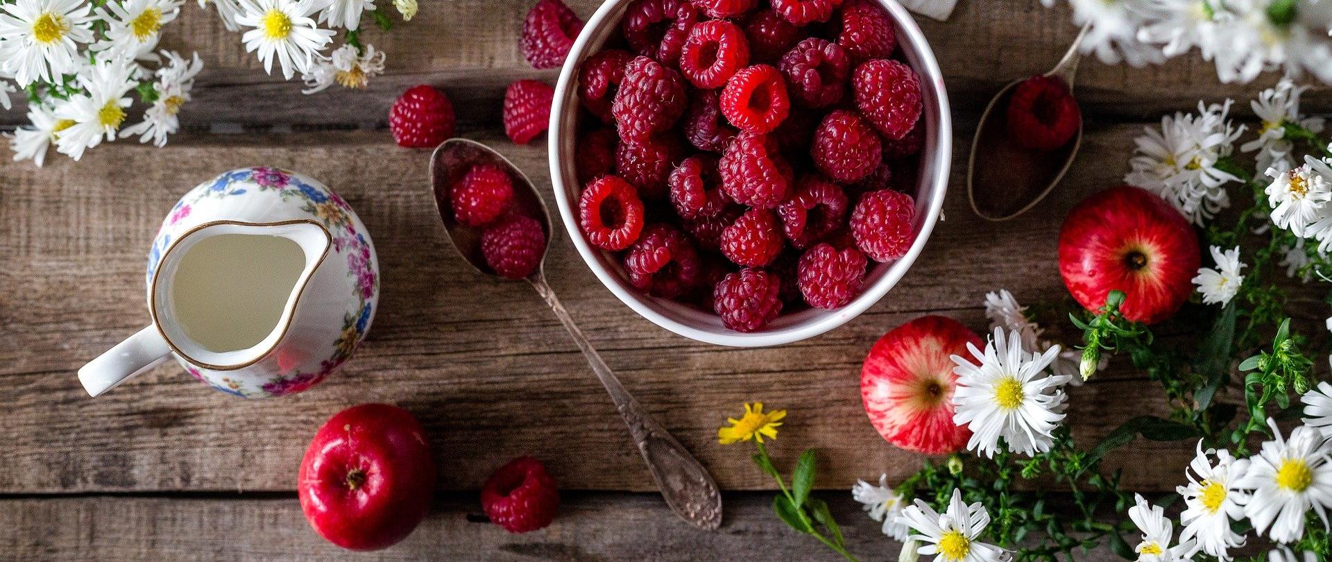 Na drewnianym stole stoi dzbanek ze śmietaną, obok leżą trzy jabłka i łyżeczka z dwoma malinami, stoi miseczka ze świeżymi malinami, a obok niej leży śieży rumianek. Na górze obok miski z malinami stoi talerzyk z herbatnikami.