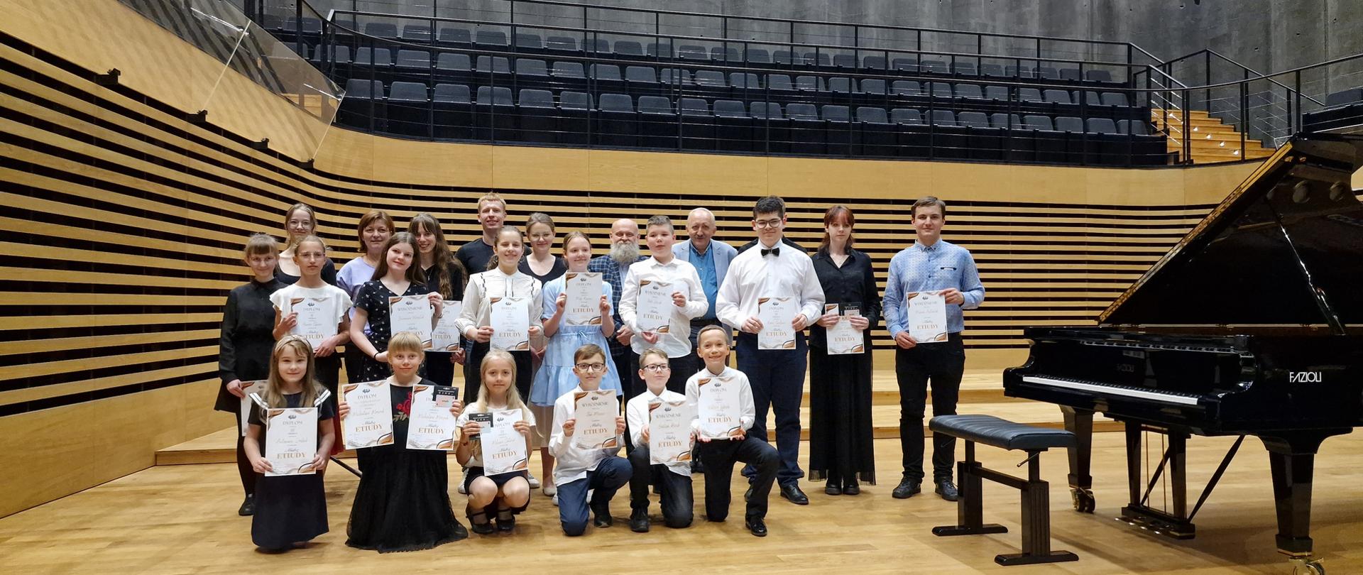 Grupa dzieci i młodzieży stoją w dwóch rzędach trzymając przed sobą dyplomy. Sześcioro dzieci w pierwszym rzędzie kuca lub klęczy. W trzecim rzędzie stoi jury.