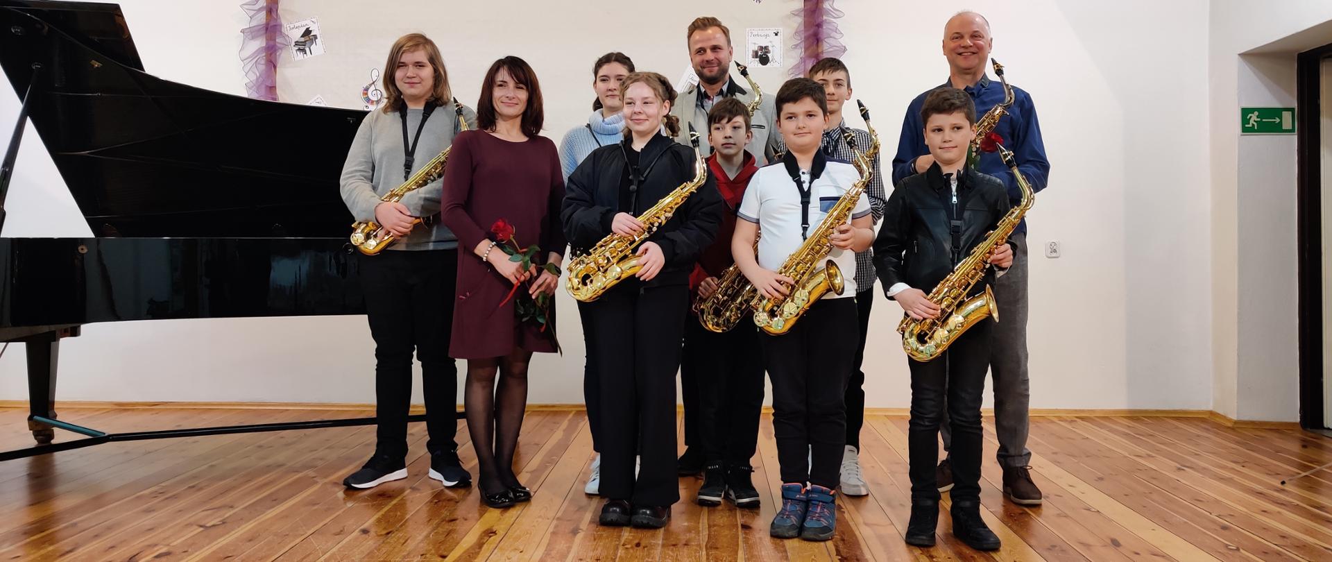 Zdjęcie grupowe podczas koncertu saksofonowego w dniu 19.04.2023 r. w auli szkoły. Na scenie klasa saksofonu oraz osoby dorosłe: od lewej strony - Halyna Hachak, Mateusz Boduch i Ryszard Żołędziewski.