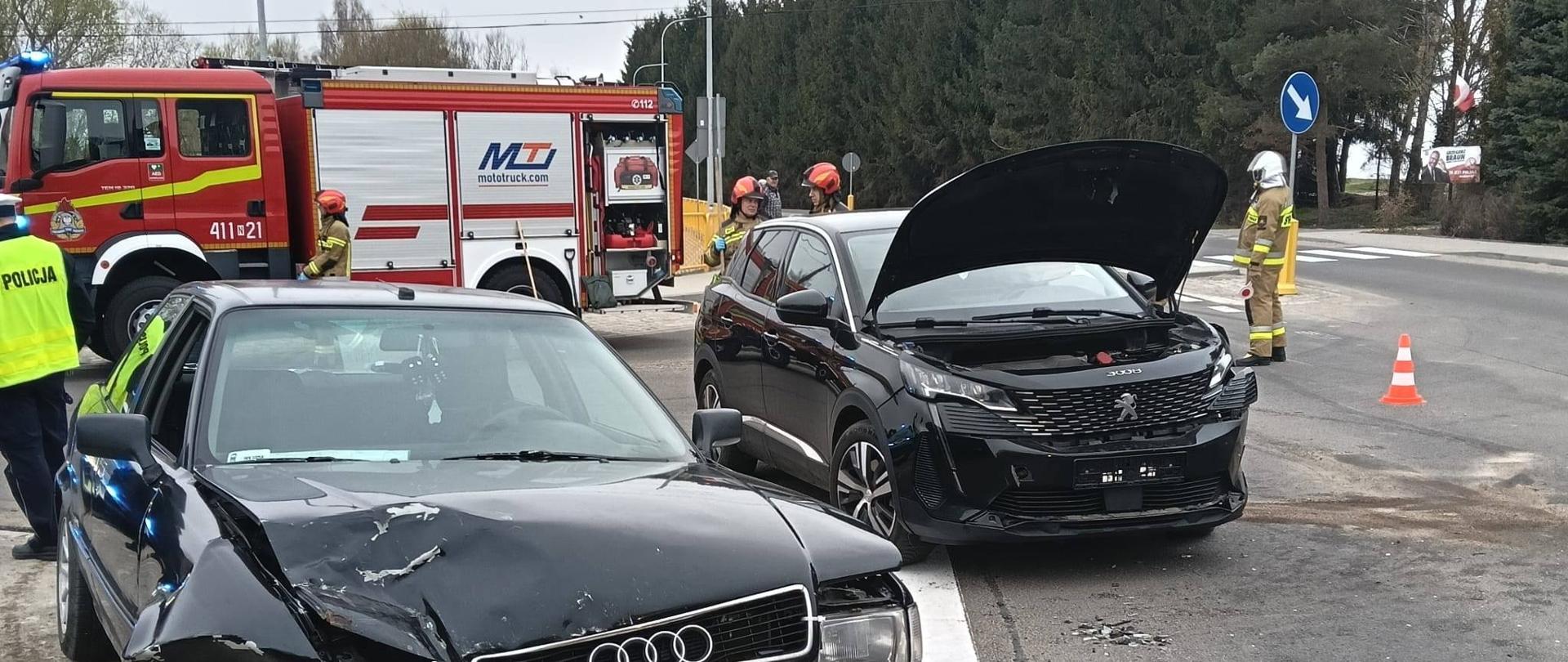 Na zdjęciu dwa uszkodzone po kolizji auta osobowe ciemnego koloru. Za nimi duży samochód strażacki i strażacy.