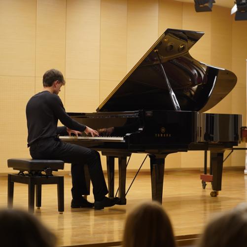Zdjęcie przedstawia innego ucznia starszej klasy grającego na fortepianie na scenie sali koncertowej, w tle baner szkoły