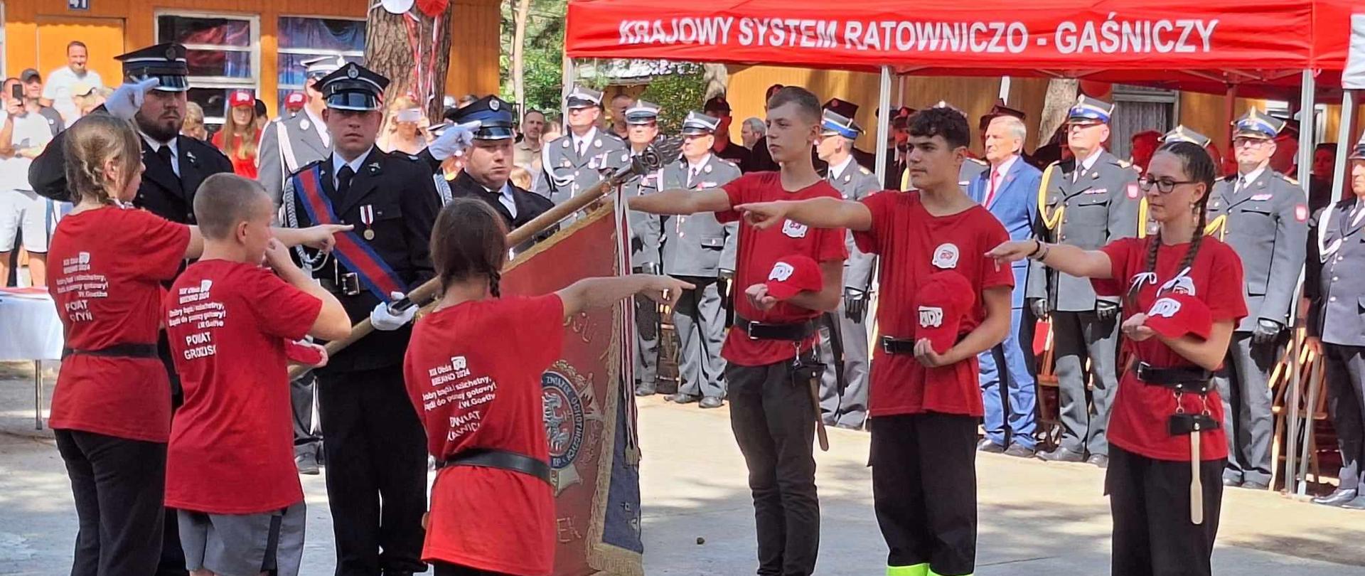 Plac apelowy. Na jego środku stoi poczet sztandarowy, a po obu stronach pochylonego sztandaru ślubowanie składa 6 obozowiczów. Każdy z nich w lewej ręce trzyma czapkę, natomiast prawe ręce mają wyciagnięte w kierunku płata sztandaru. W tle namiot w kolorze czerwonym, pod którym stoja goście - strażacy i cywile.