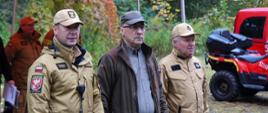 Na zdjęciu widać 3 mężczyzn, dwóch strażaków w umundurowaniu piaskowym oraz jednego w umundurowaniu leśnika. W tle widoczny jest czerwony namiot, quad, i samochód.