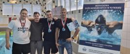 4 mężczyzn stoi wspólnie obok basenu do zdjęcia. Trzech z nich medale na szyi. Obok na sztaludze plakat zawodów.