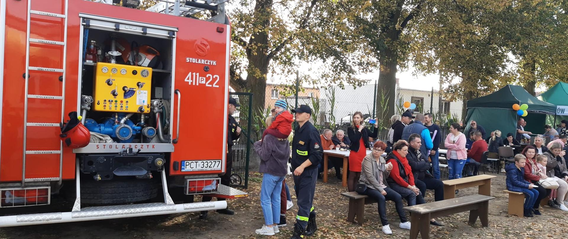 Ludzie siedzą na ławkach, obok pojazd strażacki
