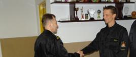 Zdjęcie wykonano wewnątrz budynku komendy powiatowej Państwowej Straży Pożarnej w pomieszczeniu świetlicy. Komendant Powiatowy Państwowej Straży Pożarnej w sępólnie krajeńskim gratuluje wyróżnionemu strażakowi ściskając mu dłoń. 