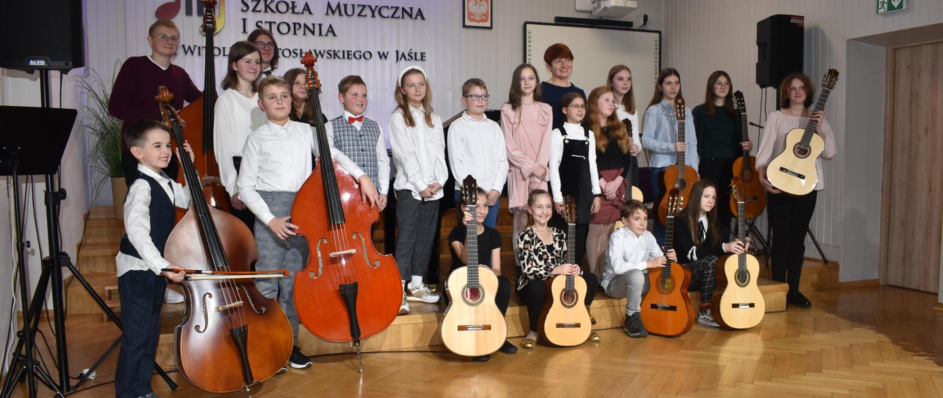 Na zdjęciu znajduje się grupa uczniów stojących na scenie w auli szkoły wraz z instrumentami, m.in. kontrabasy, gitary biorących udział w popisie, wraz z nauczycielką prowadzącą popis. W tle na ścianie od lewej strony znajduje się logo szkoły oświetlone na niebiesko, godło oraz tablica multimedialna z projektorem, kolor jasny ściany i podłoga drewniana. Po obu stronach zdjęcia znajdują się kolumny na statywach.