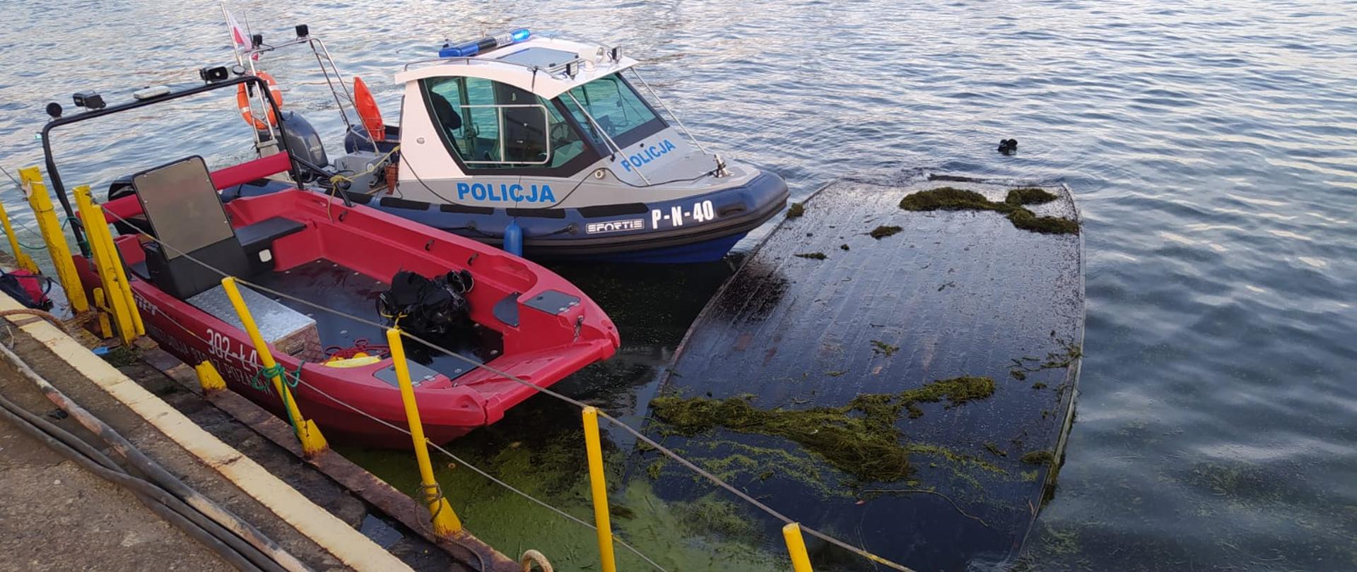 Na wodach Martwej Wisły widać przy nabrzeżu łódź wywróconą do góry dnem oraz łodzie Policji i PSP.
