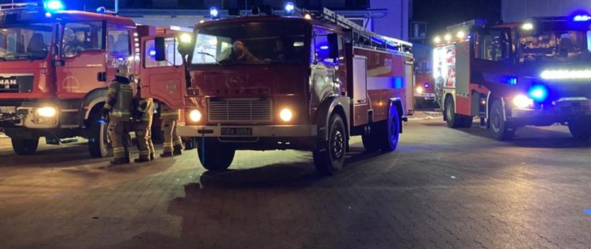 Na zdjęciu znajdują się trzy samochody ratowniczo- gaśnicze straży pożarnej ustawione przodem pojazdu . W oddali, w prawej części zdjęcia widoczny jest czwarty samochód . Obok pojazdu znajdującego się w lewej części zdjęcia znajduje się trzech strażaków. 