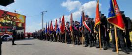poczty sztandarowe stojące w szeregu w czasie uroczystości w tle budynek remizy z muralem