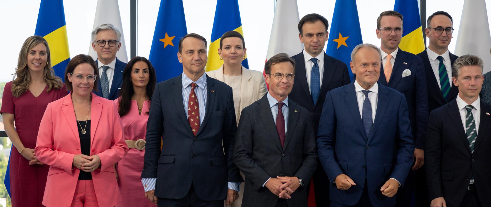 Grupa dwunastu uczestników spotkania pozuje do wspólnego zdjęcia na tle naprzemiennie ustawionych flag Szwecji, Polski i Unii Europejskiej oraz przeszklonej ściany z widokiem na zieleń. W pierwszym rzędzie widać Wicepremiera Sikorskiego oraz Premierów Szwecji i Polski Ulfa Kristerssona i Donalda Tuska. W tylnym rzędzie stoi wiceminister MSZ Artur Harazim. Zdjęcie ma charakter oficjalny i dyplomatyczny.