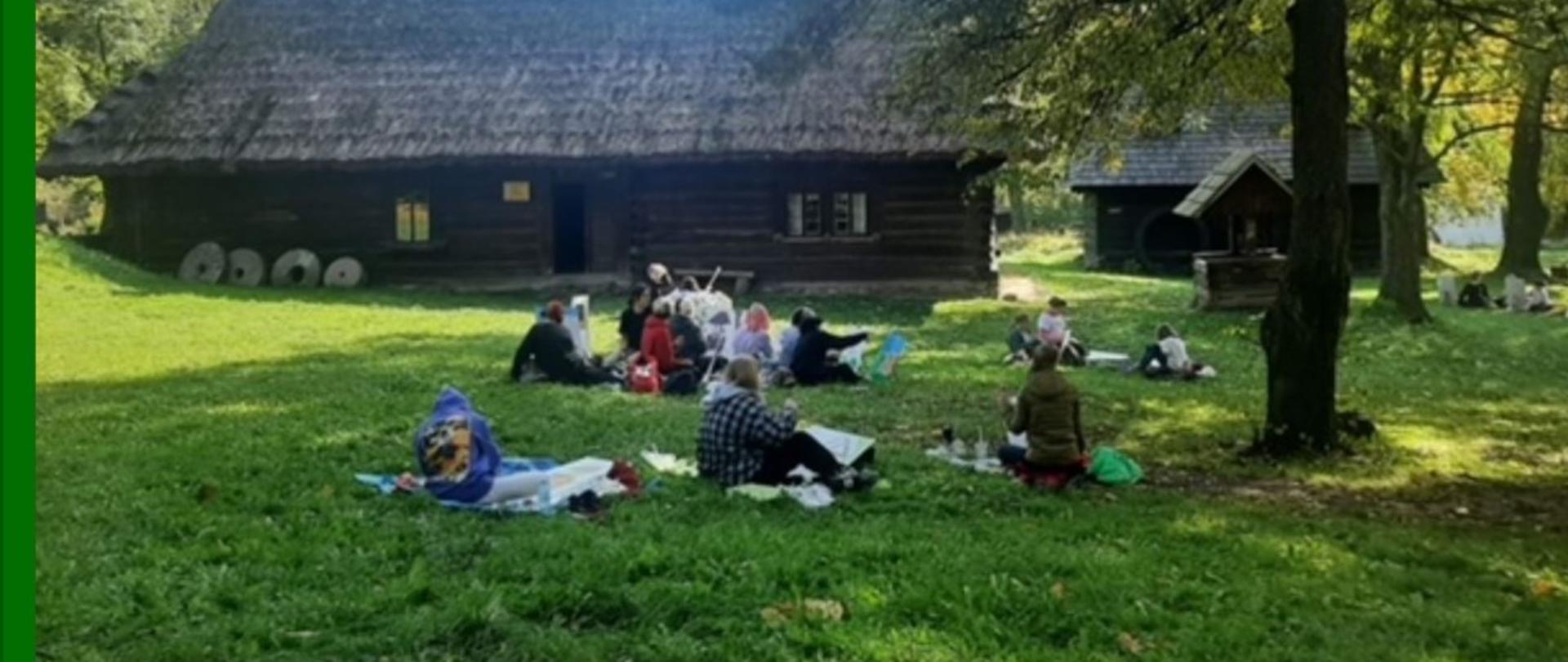 Skansen - osoby malujące pejzaż, w tle drewniane zabudowania.