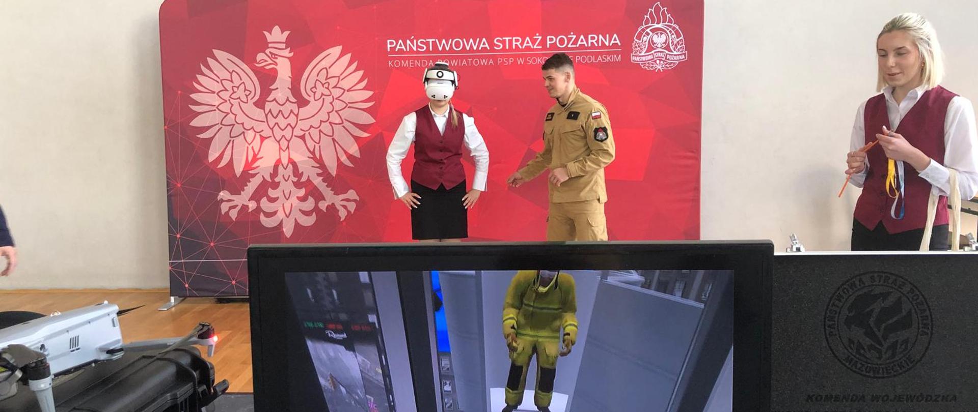 Zdjęcie przestawia funkcjonariusza PSP w ubraniu koszarowym koloru piaskowego. Obok niego w czarnej spódnicy, białej koszuli i czerwonej kamizelce stoi kobieta z okularami VR. Oboje stoją na tle bilbordu KP PSP Sokołów Podlaski koloru czerwonego. W bliższym planie widzimy monitor telewizyjny z programem szkoleniowym ze strażakiem.