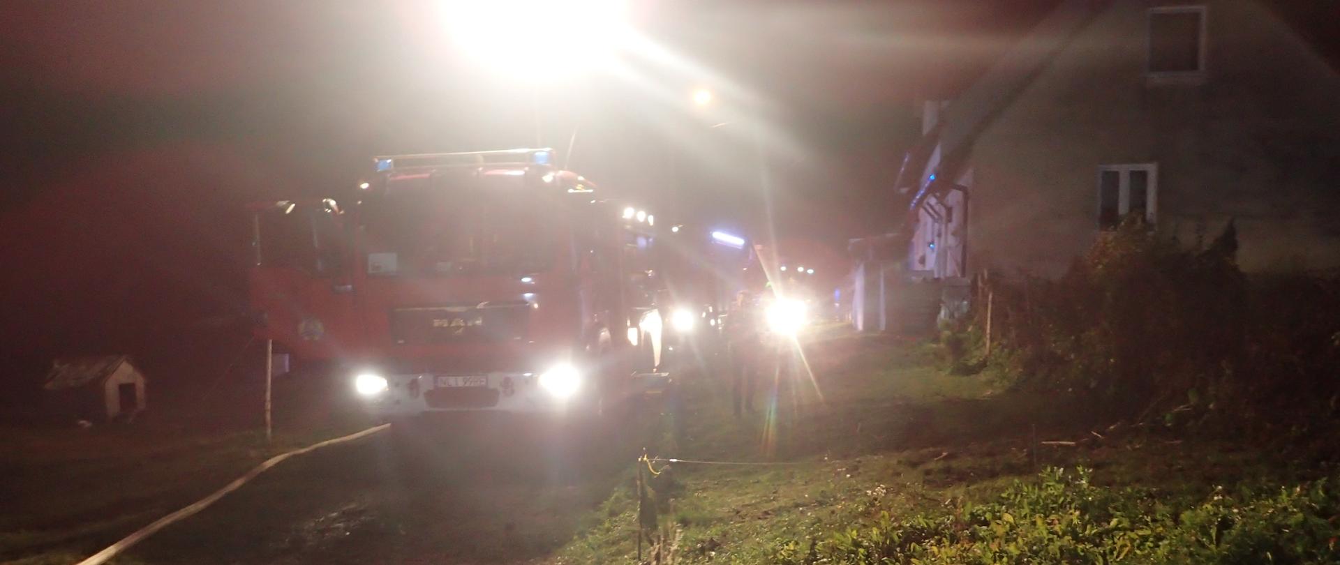 Zdjęcie przedstawia ustawione pojazdy pożarnicze równolegle do zagrożonego budynku. Pora nocna