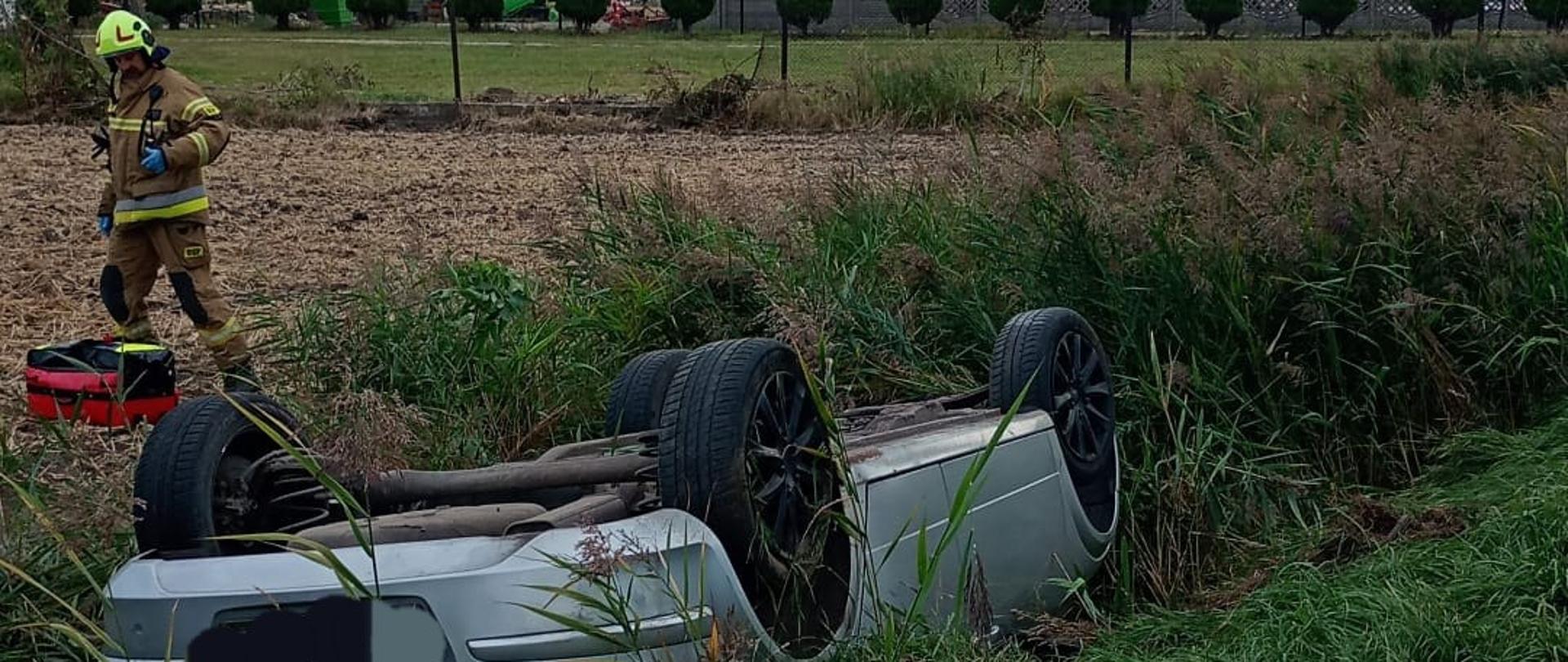 Na zdjęciu widać samochód osobowy, który dachował i leży do góry kołami w przydrożnym rowie. Auto jest srebrnego koloru, a jego podwozie i koła są skierowane do góry. Obok samochodu stoi strażak w pełnym umundurowaniu ochronnym i hełmie, trzymający sprzęt ratowniczy. W tle widać pole, drzewa, zabudowania gospodarcze oraz silosy zbożowe. 