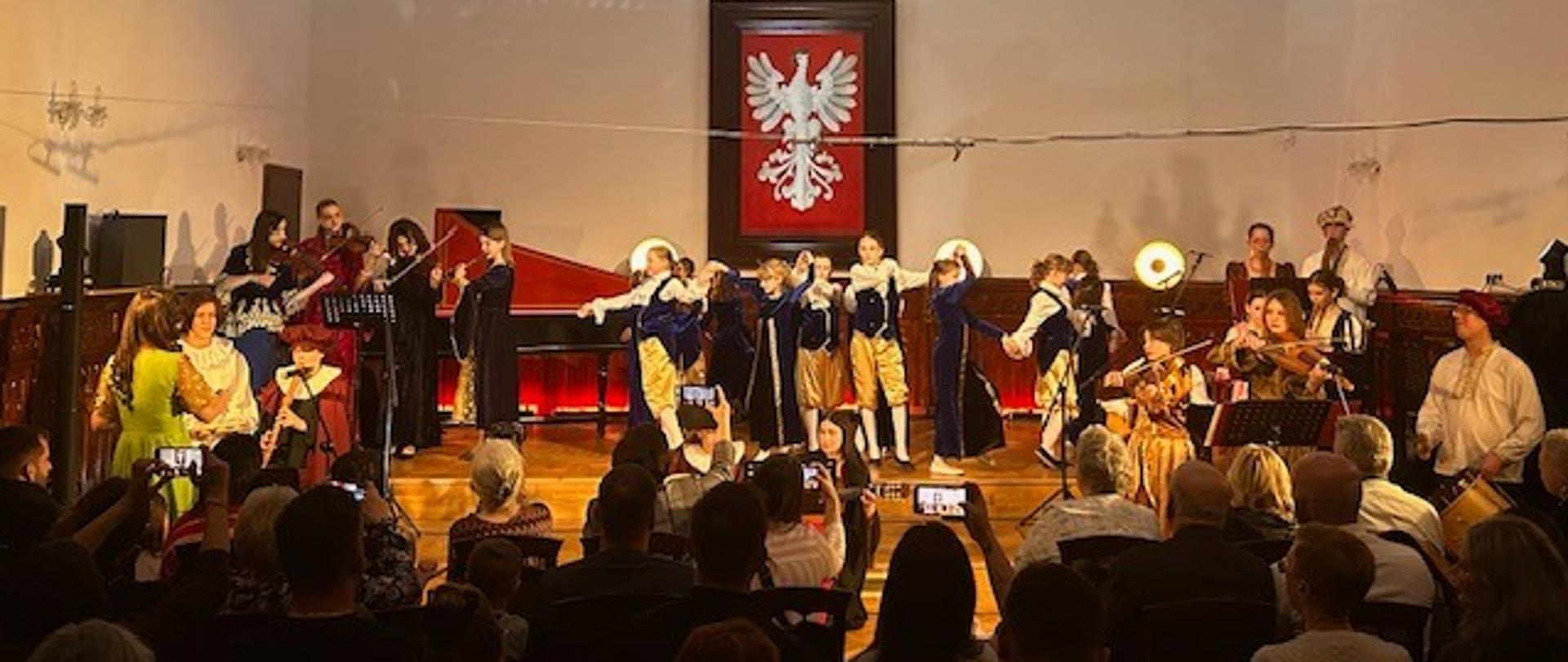 Występ zespołów muzyki dawnej i grupy tanecznej na scenie w Sali Królewskiej podczas koncertu z repertuarem historycznym, z udziałem publiczności.