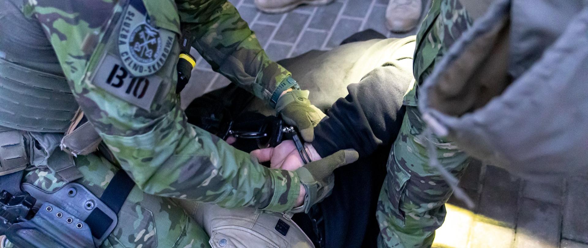 Funkcjonariusze w umundurowaniu obezwładniają osobę leżącą na chodniku i zakładają kajdanki.