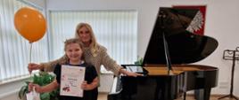 Dziewczynka stoi obok fortepianu, trzyma dyplom i upominek, za nią stoi kobieta trzymająca balonik.