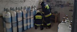 W budynku technicznym dwóch strażaków zabezpiecza w pozycji stojącej przeniesioną butle z tlenem