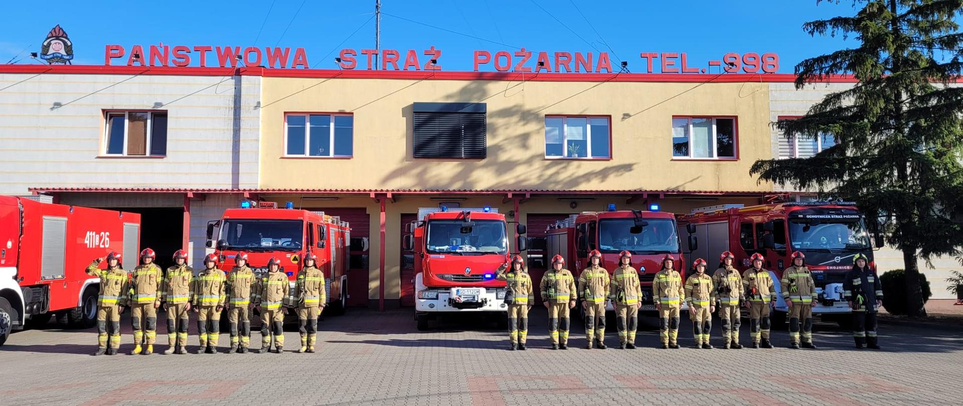 Siedemnastu strażaków ustawionych w szeregu w ubraniach specjalnych. Szesnastu strażaków w czerwonych hełmach na głowie jeden stojący z lewej strony w rażącym zielonym, dowódcy zmian oddają honory. Strażacy ustawieni na tle pięciu czerwonych samochodów z włączoną sygnalizacją świetlą, samochody ustawione w bramach garażowych Komendy Państwowej Straży Pożarnej w Chojnicach.