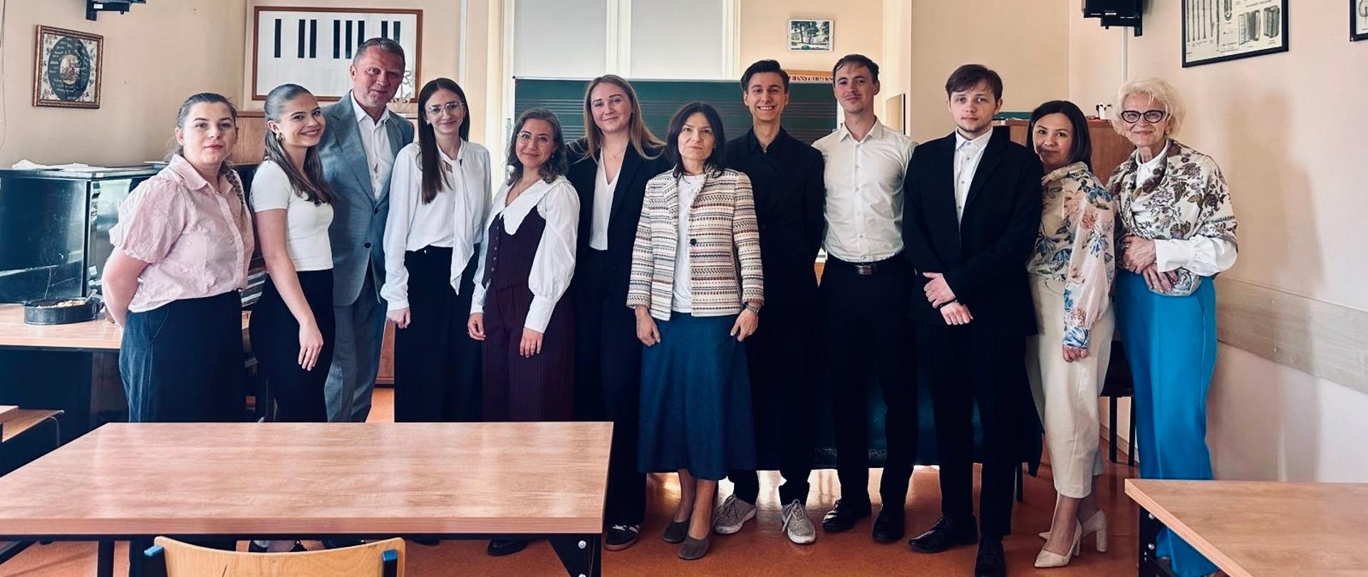 Grupowe zdjęcie dwunastu osób stojących w rzędzie w sali lekcyjnej muzycznej. W centrum znajduje się kobieta w jasnej marynarce w paski, otoczona przez osoby ubrane w eleganckie stroje - białe koszule, czarne marynarki i spódnice. Po lewej stronie widoczna jest tablica z klawiaturą pianina, a na ścianach wiszą głośniki i oprawione plakaty. Pomieszczenie ma jasnobeżowe ściany, sufit z panelami i lampami fluorescencyjnymi. Na pierwszym planie widoczne są drewniane stoły.