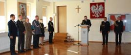 Na zdjęciu strażacy stoją w mundurach wyjściowych, jedna osoba przemawia za mównicą, po lewej stronie kilka osób stoi w ubraniu cywilnym na ścianie wisi godło Polski, Krzyż i sztandar.