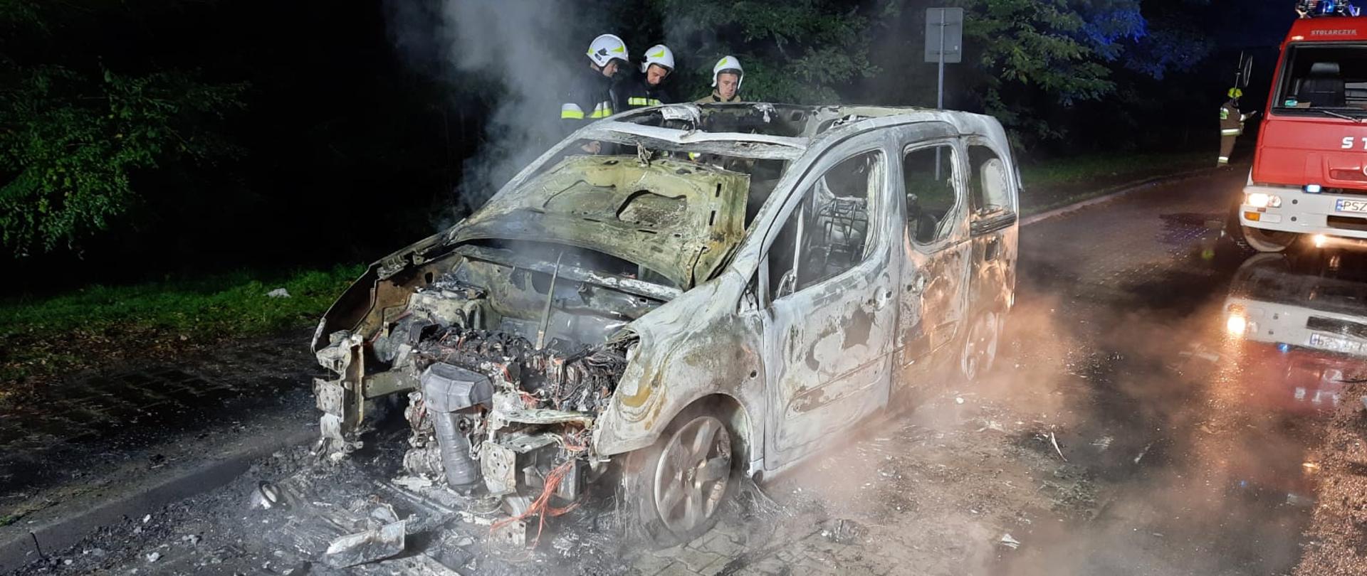 Widać spalony samochód na drodze, jest noc, z tyłu pojazd straży pożarnej