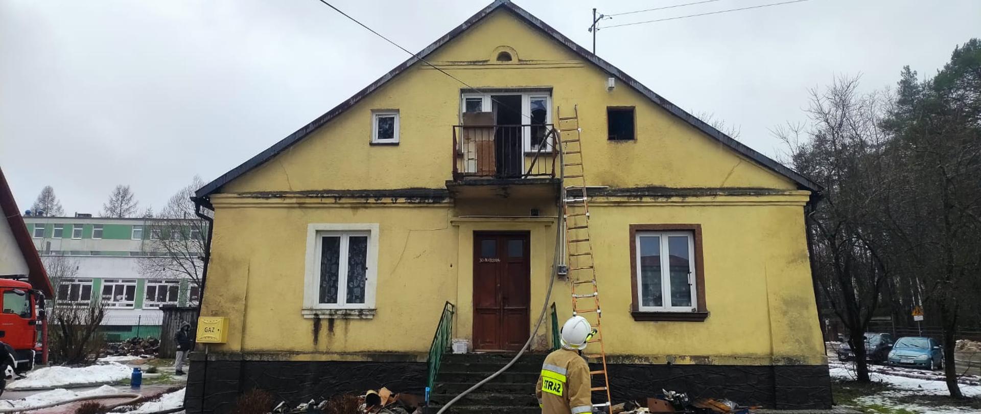 Zdjęcie przedstawia budynek mieszkalny gdzie doszło do pożaru na poddaszu. Do balkonu przystawiona została drabina, widać też wąż gaśniczy wciągnięty do wnętrza domu. Przed budynkiem stoi strażak.