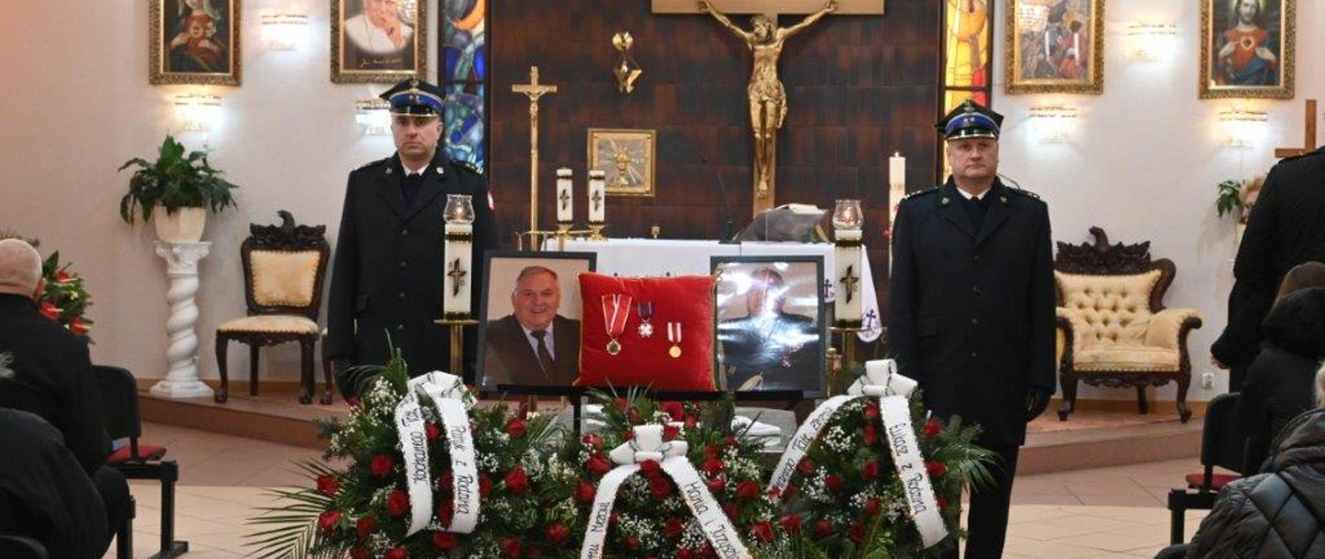 Dwóch strażaków stoi na warcie w kaplicy domu pogrzebowego. Po lewej stronie stoi Komendant Powiatowy Państwowej Straży Pożarnej w Tucholi. Komendant ma na sobie granatowy mundur wyjściowy. Po prawej stronie stoi Zastępca Komendanta Powiatowego Państwowej Straży Pożarnej w Tucholi. Zastępca Komendanta ma na sobie granatowy mundur wyjściowy. Na środku widać wieńce pogrzebowe. Nad wieńcami pogrzebowymi widać na środku czerwoną poduszkę z odznaczeniami. Po prawej i lewej stronie poduszki widnieją zdjęcia zmarłego komendanta oraz świece. Po prawej i lewej stronie widać krzesła i siedzących ludzi. W tle widać ołtarz oraz wiszący duży krzyż. Ściana za ołtarzem jest koloru czarno-brązowego. Na ołtarzu widać biały obrus, świece oraz mniejszy krzyż. Po prawej i lewej stronie od ołtarza wiszą na ścianie święte obrazy, stoją fotele. Podłoga jest koloru białego. Ściany są koloru białego. 