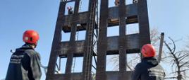 Dwóch egzaminatorzy w czerwonych kaskach stojący pod wspinalnią strażacką. Na wspinalni trzech ratowników przygotowujących się do zjazdu na linie