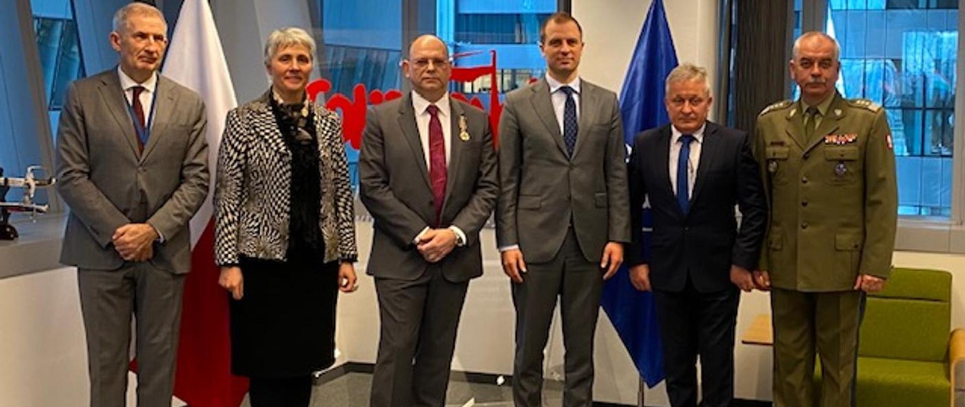 Odznaczony mężczyzna stoi na tle logo Solidarności w towarzystwie Ambasadora i gości