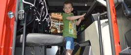 Wnętrze kabiny samochodu ratowniczo-gaśniczego. W niej stoi chłopczyk w zielonej koszulce i jeansowych krótkich spodniach. 