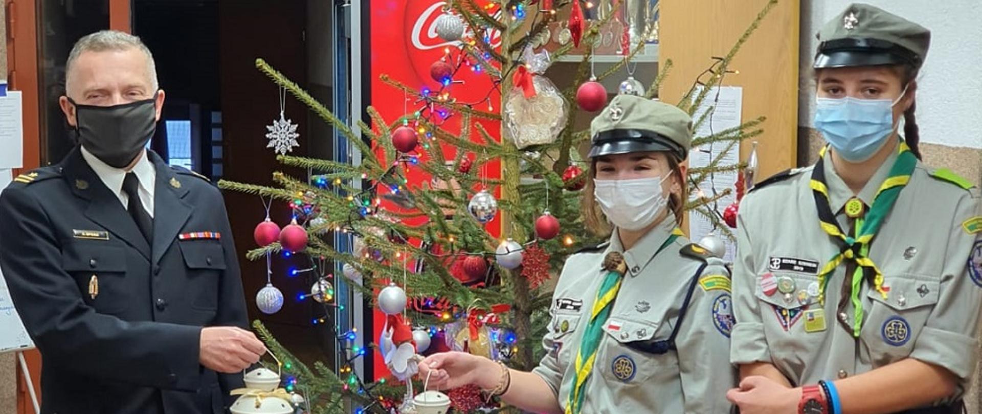 Zdjęcie zrobione w budynku Komendy Powiatowej Państwowej Straży Pożarnej w Będzinie. Po lewej stronie stoi Komendant Powiatowy mł. bryg. Arkadiusz Spera ubrany w mundur wyjściowy, po prawej stronie stoją dwie dziewczyny ubrane w mundury harcerskie. Komendant i jedna z harcerek trzymają w ręku lampion z Betlejemskim Światełkiem Pokoju. W tle stoi choinka z kolorowymi ozdobami i świecącymi lampkami
