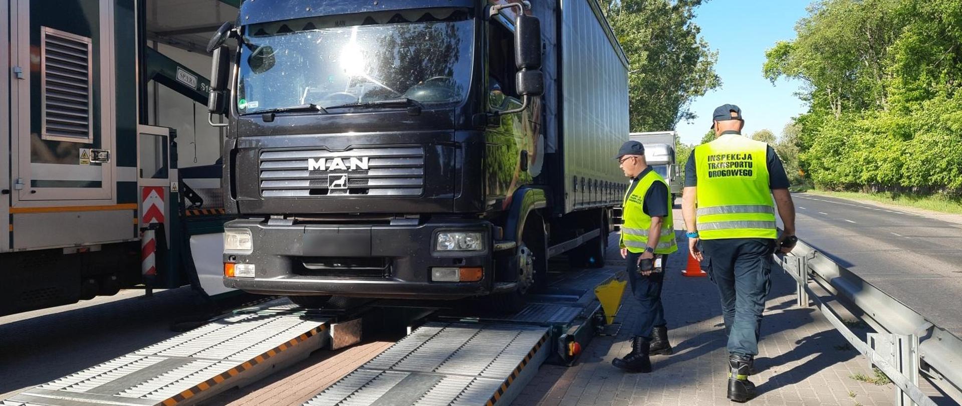 Inspektorzy podczas kontroli z wykorzystaniem Mobilnej Jednostki Diagnostycznej, na matach najazdowych stojący czarny pojazd.