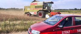 Samochód operacyjny straży pożarnej stoi na polu. Na drugim planie trwa młócenie zboża.