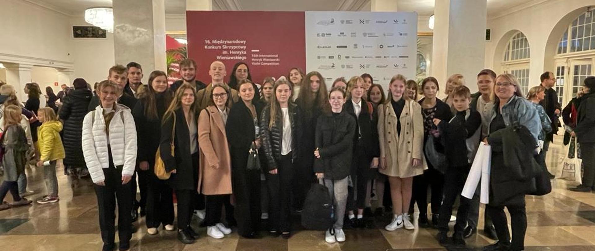 Uczniowie stoją holu Filharmonii, w tle logo 16 Międzynarodowego Konkursu Skrzypcowego im. H. Wieniawskiego