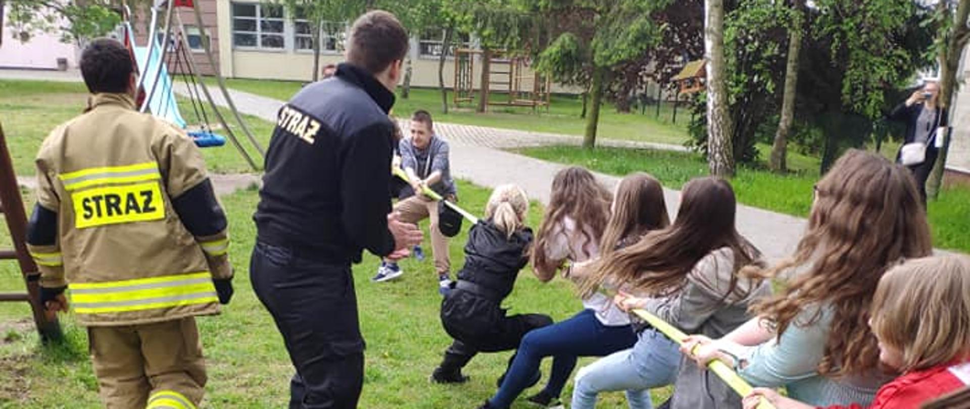 Na zdjęciu grupa dzieci na trawie, podczas konkurencji w przeciąganiu strażackiego węża. W roli sędziów dwóch strażaków. W tle drzewa, budynek szkoły oraz elementy infrastruktury placu zabaw: huśtawka i ślizgawka.