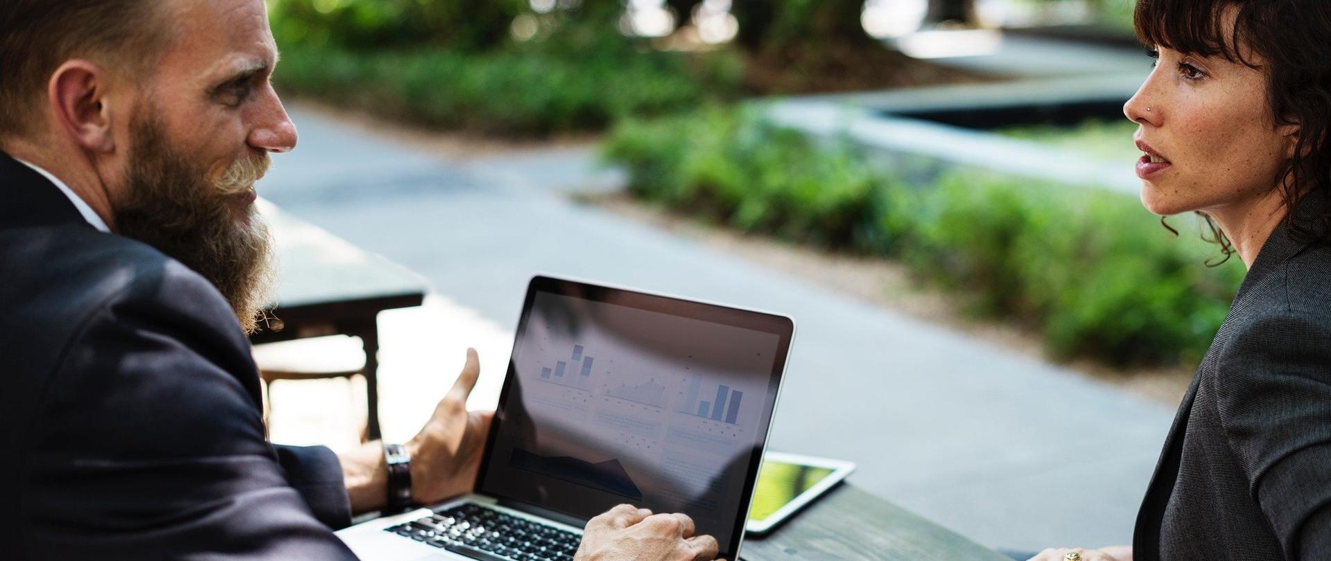 Mężczyzna w garniturze siedzi przy stole i rozmawia z kobietą. Mężczyzna ma przed sobą otwartego laptopa.